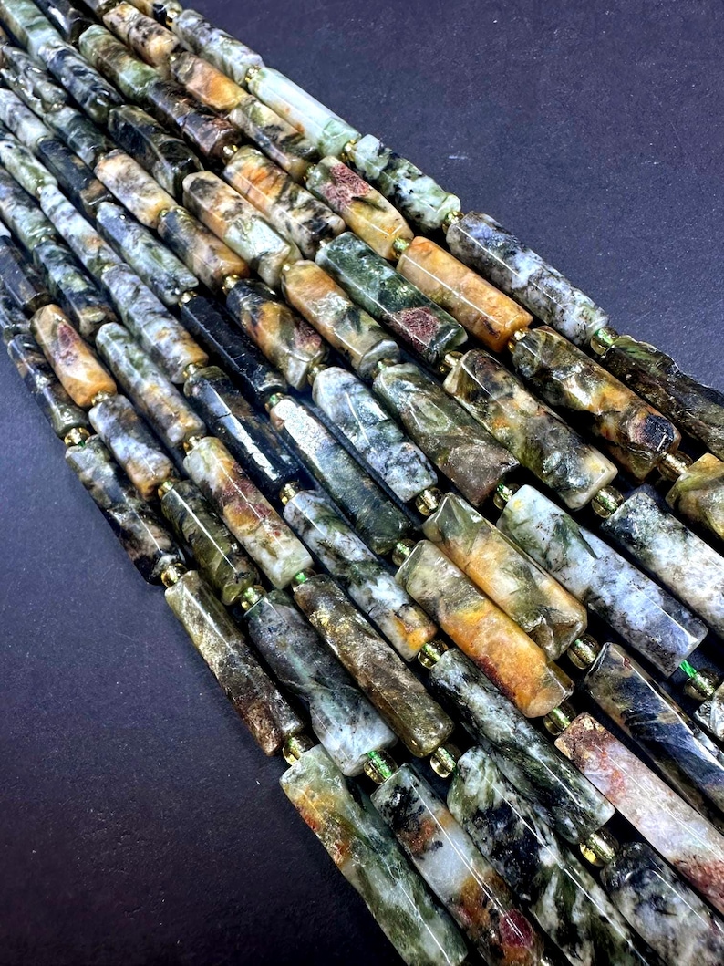 May include: A collection of cylindrical beads arranged in parallel strands. The beads feature a marbled pattern with variegated shades of forest green, earthy brown, cream, and black, with a polished finish and small gold-toned spacers.