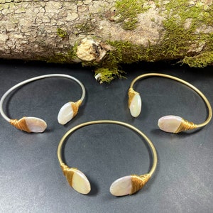 Pu&ograve; includere: Tre braccialetti in oro e argento con un accento di conchiglia bianca. I braccialetti sono su una superficie di legno con muschio.