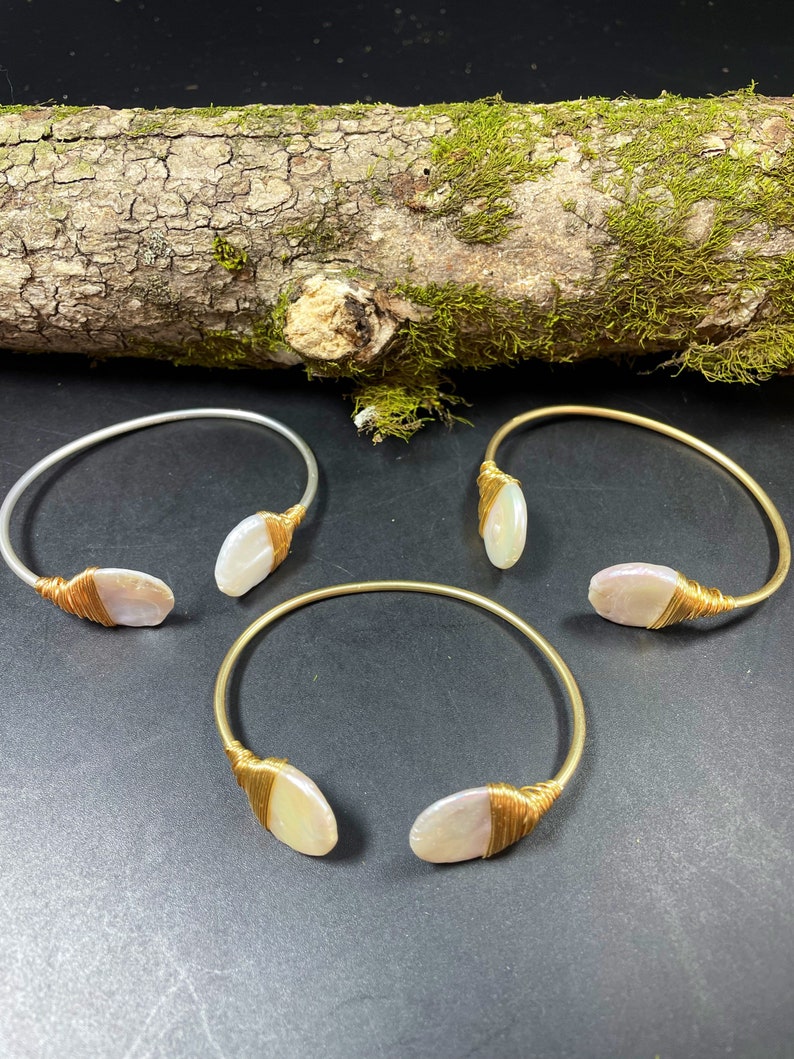 Pu&ograve; includere: Tre braccialetti in oro e argento con un accento di conchiglia bianca. I braccialetti sono su una superficie di legno con muschio.
