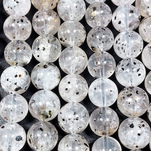 May include: A close-up shot of multiple clear quartz beads with black and brown inclusions. The beads are spherical and appear to be strung together. The beads are arranged in a grid pattern, filling the frame.