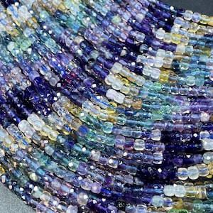 May include: Close-up of strands of faceted beads in a variety of colors, including purple, blue, green, and yellow. The beads are small and square-shaped, creating a textured surface. The beads are likely for jewelry making.
