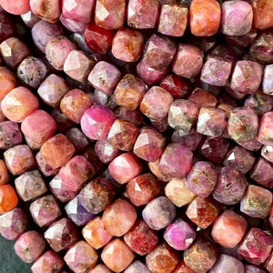 May include: Close-up of a collection of faceted gemstone beads in shades of pink, red, and purple. The beads have a square shape with multiple facets, creating a sparkling effect. The gemstones are strung together, suggesting they are for jewellery making.