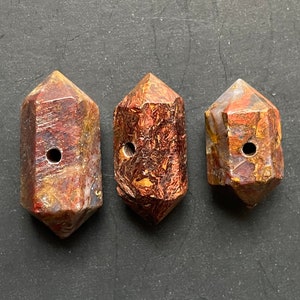 May include: Three brown, red and gold gemstone beads with a hole drilled through the centre. The beads are shaped like a long, narrow, six-sided crystal.