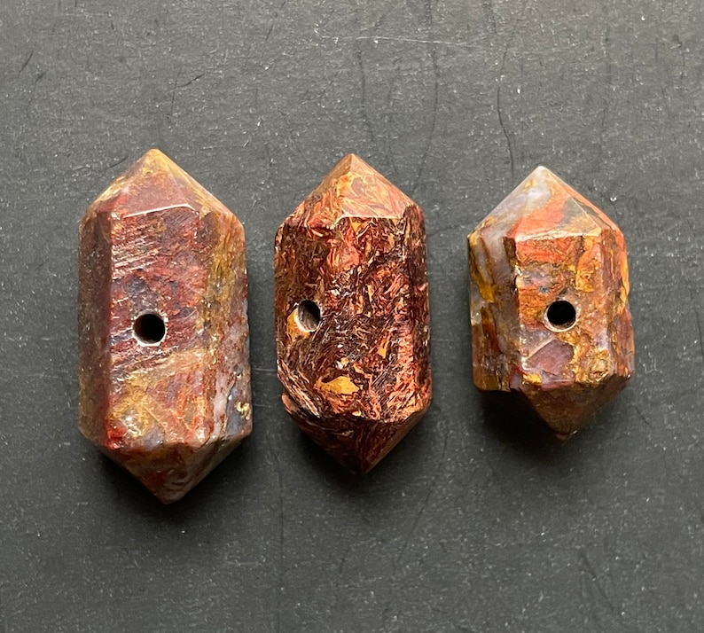 May include: Three brown, red and gold gemstone beads with a hole drilled through the centre. The beads are shaped like a long, narrow, six-sided crystal.