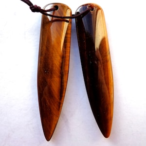 May include: Two polished tiger's eye pendants with a brown cord. The pendants are elongated, with a pointed bottom and a rounded top. The color is a mix of brown and gold, with visible striations. The pendants are likely for jewelry.