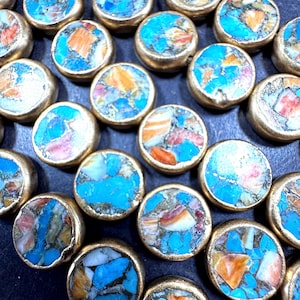 May include: Close-up of a collection of round, gold-colored jewelry components. Each component features a mosaic of turquoise, orange, and cream-colored stones. The components are arranged closely together, creating a textured surface.