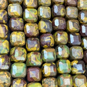 Puede incluir: Primer plano de una hebra de cuentas de piedras preciosas. Las cuentas son una mezcla de colores verde, marrón y amarillo con un patrón moteado. Las cuentas son facetadas y tienen un acabado pulido.