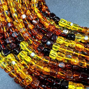 May include: Close-up of strands of amber-colored beads. The beads are square-shaped and vary in color from light yellow to dark brown. The beads are strung together, creating a textured and layered effect. The background is a dark gray.