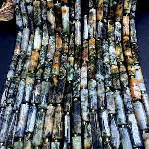 May include: Close-up of a strand of gemstone beads. The beads are rectangular with a mix of colors including green, brown, black, and cream. Each bead is separated by a small gold-colored spacer. The beads are strung together, creating a textured surface.