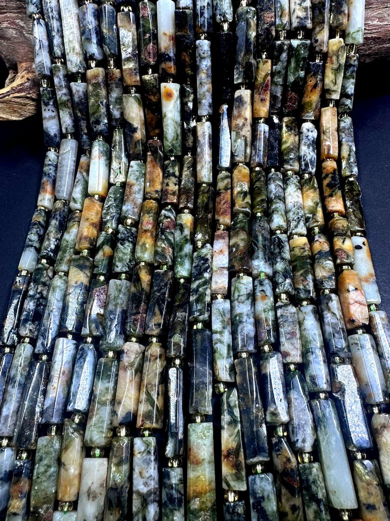 May include: Close-up of a strand of gemstone beads. The beads are rectangular with a mix of colors including green, brown, black, and cream. Each bead is separated by a small gold-colored spacer. The beads are strung together, creating a textured surface.