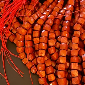 May include: A strand of red carnelian beads, each bead is faceted and has a square shape. The beads are strung on a red thread.