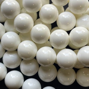 May include: A strand of white round beads, made of polished ceramic or porcelain. The beads are smooth and glossy, with a slight variation in size and shape.