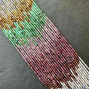 May include: A variety of gemstone beads in different colors, including green, pink, brown, and white. The beads are arranged in rows on a dark gray surface.