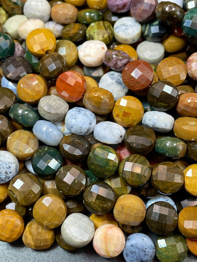 May include: A close-up of a collection of round, faceted gemstone beads in various colors, including green, brown, yellow, white, and red. The beads are made of natural stone and have a polished finish.
