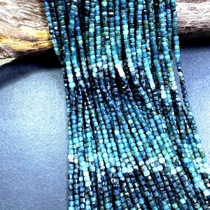 May include: Strands of small, square-cut beads in shades of teal, turquoise, and navy blue. The beads are strung together, creating a cascading effect. The background includes a piece of wood and a dark blue surface.