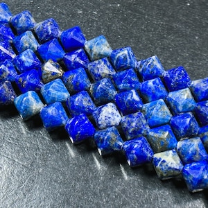 Puede incluir: Primer plano de un hilo de cuentas de lapislázuli facetadas, de corte cuadrado. Las cuentas son de un azul profundo y rico con motas doradas y blancas, dispuestas en filas diagonales. Las cuentas miden aproximadamente 0,8 cm.