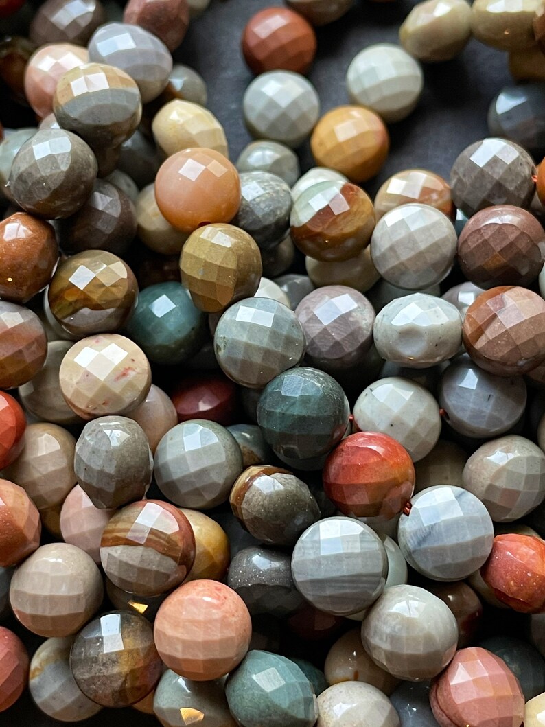 May include: A close-up of a collection of colourful, faceted gemstone beads. The beads are a mix of brown, grey, green, and red tones. The beads are all round and have a smooth, polished finish.
