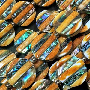 May include: Close-up of multiple round beads with a striped design. The beads feature a combination of light brown wood and iridescent abalone shell. Each bead is connected with a small silver-colored bead, creating a pattern of repeating circles.