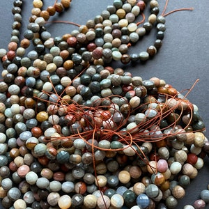 May include: A strand of multi-coloured gemstone beads, featuring a variety of shades of brown, grey, and red. The beads are faceted and have a smooth, polished finish.