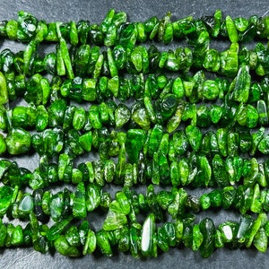 Puede incluir: Primer plano de hebras de virutas de piedras preciosas de color verde vibrante. Las piedras pulidas varían en tamaño y forma, creando una apariencia texturizada. Las cuentas están ensartadas, listas para la fabricación de joyas u otras manualidades.