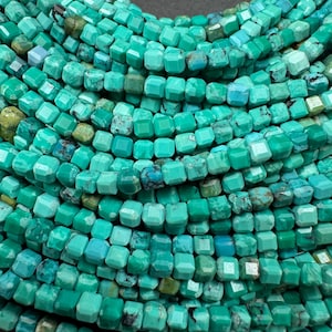 May include: Close-up of a turquoise necklace. The necklace features multiple strands of faceted, square-shaped beads in varying shades of turquoise and blue-green. The beads are strung together, creating a layered effect. The background is dark.