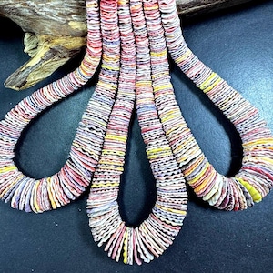 May include: Close-up of several strands of colorful, handcrafted shell necklaces. The necklaces feature a variety of colors including red, yellow, white, and purple. The background includes a piece of wood and a dark surface.