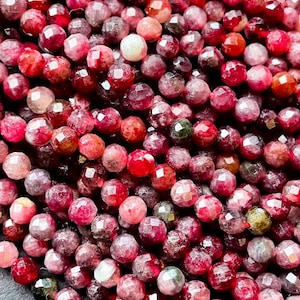 May include: A strand of faceted red gemstone beads. The beads are a mix of pink, red, and green hues. The beads are round and have a smooth, polished finish.
