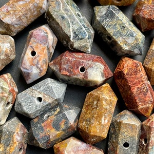 May include: A collection of 18 irregularly shaped, multi-coloured, polished gemstone beads with a hole drilled through the centre of each bead. The beads are a mix of brown, red, grey, and black with white and gold accents.
