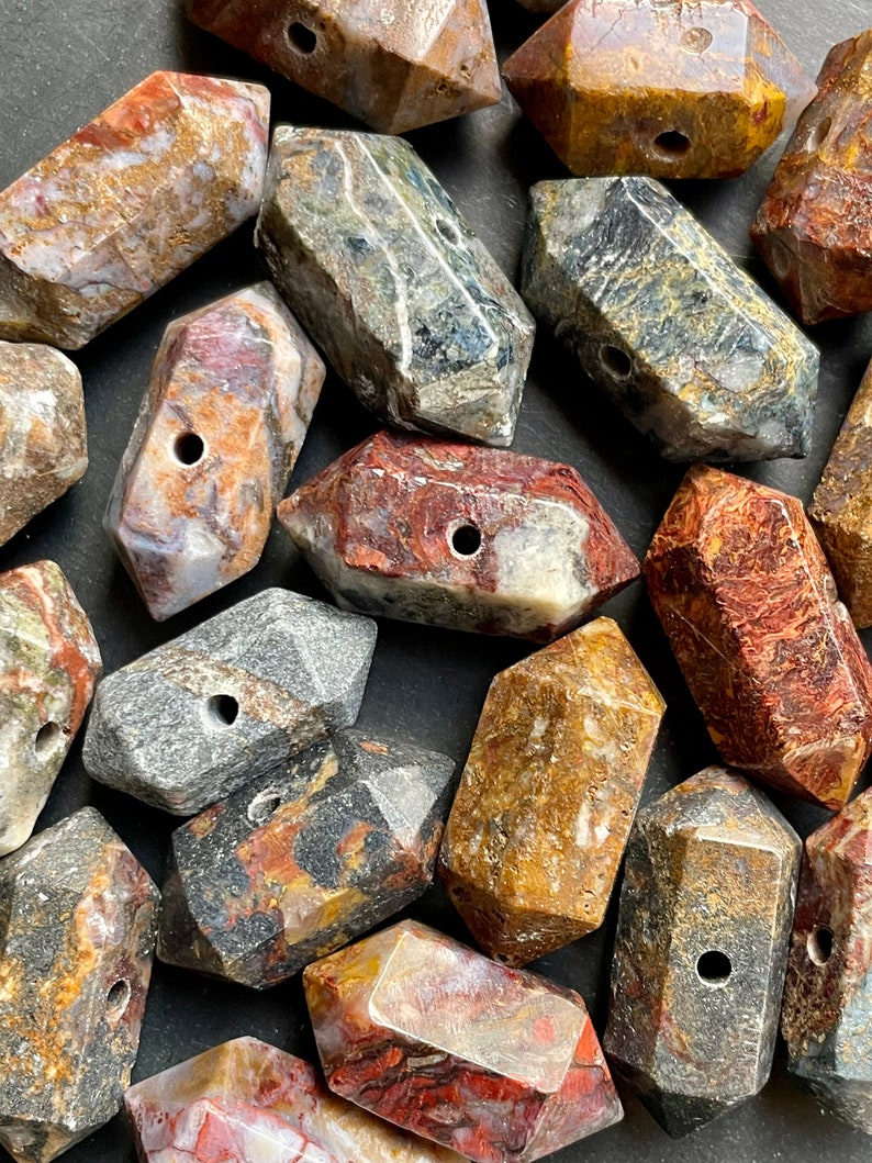 May include: A collection of 18 irregularly shaped, multi-coloured, polished gemstone beads with a hole drilled through the centre of each bead. The beads are a mix of brown, red, grey, and black with white and gold accents.