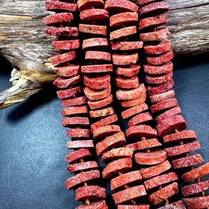 May include: Strands of red coral beads, each a flat, irregular disc shape. The beads are strung together, showcasing their natural texture and color variations. The background includes a piece of driftwood.