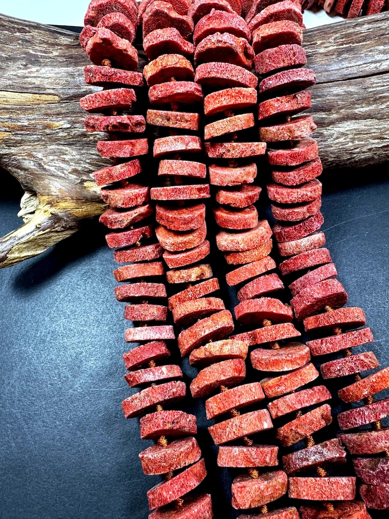 May include: Strands of red coral beads, each a flat, irregular disc shape. The beads are strung together, showcasing their natural texture and color variations. The background includes a piece of driftwood.