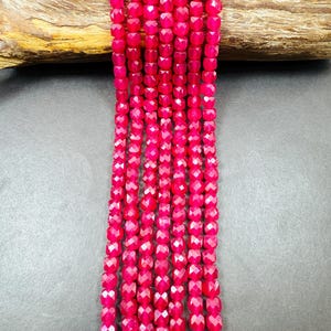 May include: Strands of faceted, hot pink beads. The beads are cube-shaped with multiple facets, creating a sparkling effect. The beads are strung together, forming vertical lines. The background includes a piece of wood and a dark gray surface.