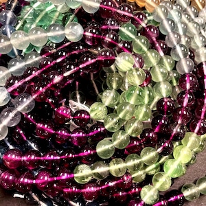 May include: A close-up of a pile of round, smooth, gemstone beads in various shades of green, purple, and blue. The beads are arranged in a circular pattern, creating a visually appealing display.