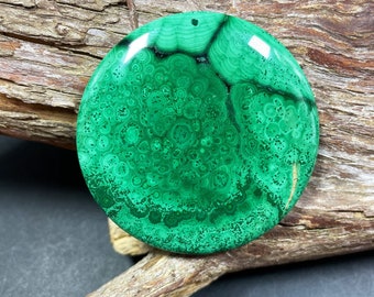 May include: A circular malachite pendant, displaying a rich green hue and complex, swirling designs. The stone measures approximately 5 cm in diameter and features a small hole for threading. It is presented on a piece of weathered wood.