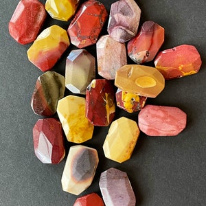 May include: A collection of 17 polished, faceted, red, yellow, and purple jasper stones. The stones are in various shapes and sizes, and they are arranged in a random pattern.