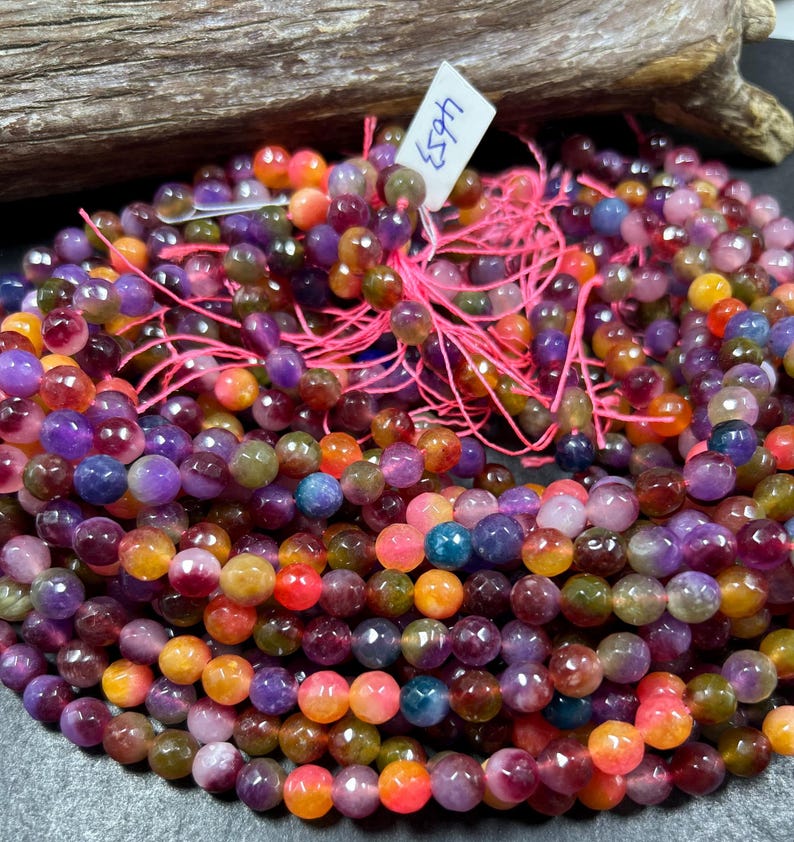 May include: A strand of multi-colored faceted gemstone beads. The beads are a mix of purple, pink, orange, and yellow. The beads are strung on a pink thread.