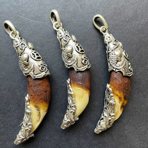 Puede incluir: Tres colgantes con diseño de diente natural. Cada diente tiene una ornamentada montura de metal plateado y un lazo para colgar. Los dientes hacen la transición de un marrón oscuro a un blanco cremoso.