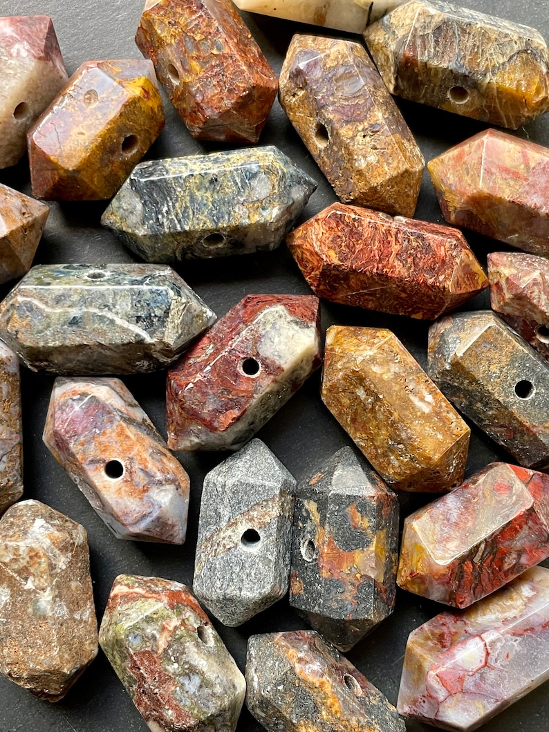 May include: A collection of 18 irregularly shaped, multi-coloured, polished stone beads with a hole drilled through the centre. The beads are a mix of brown, red, grey, and black with swirling patterns.