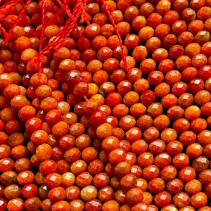 K&ouml;nnte beinhalten: Eine Str&auml;hne aus orangefarbenen Korallenperlen, jede Perle ist facettiert und hat eine strukturierte Oberfl&auml;che. Die Perlen sind auf einer roten Schnur aufgef&auml;delt.