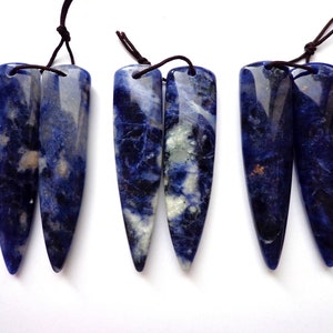 May include: Six blue sodalite pendants with dark brown cords. The pendants are rectangular with a pointed bottom and a hole at the top for the cord. The sodalite has a mix of dark blue, white, and gray colors.