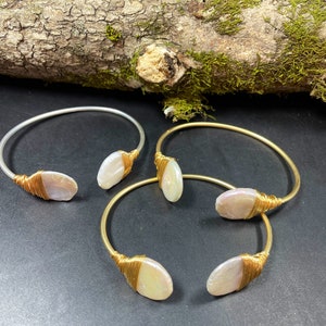 Pu&ograve; includere: Tre braccialetti avvolti in filo d'oro con accenti di perla ovale bianca. I braccialetti sono su una superficie grigio scuro.