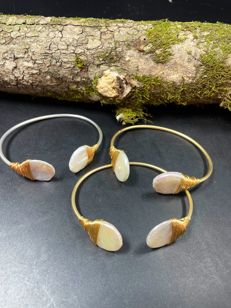 Pu&ograve; includere: Tre braccialetti avvolti in filo d'oro con accenti di perla ovale bianca. I braccialetti sono su una superficie grigio scuro.