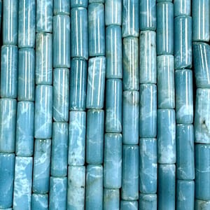 Peut inclure: Gros plan sur une collection de perles cylindriques bleu clair. Les perles présentent un aspect marbré avec des veines blanches et bleu foncé. Elles sont disposées en grille, créant une surface texturée. Ces perles sont adaptées à la création de bijoux.