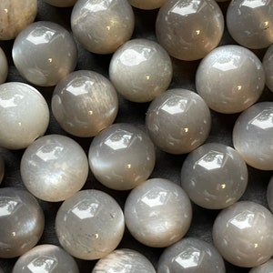 May include: A collection of smooth, round, grey moonstone beads. The beads have a subtle shimmer and are arranged in a close-up view.