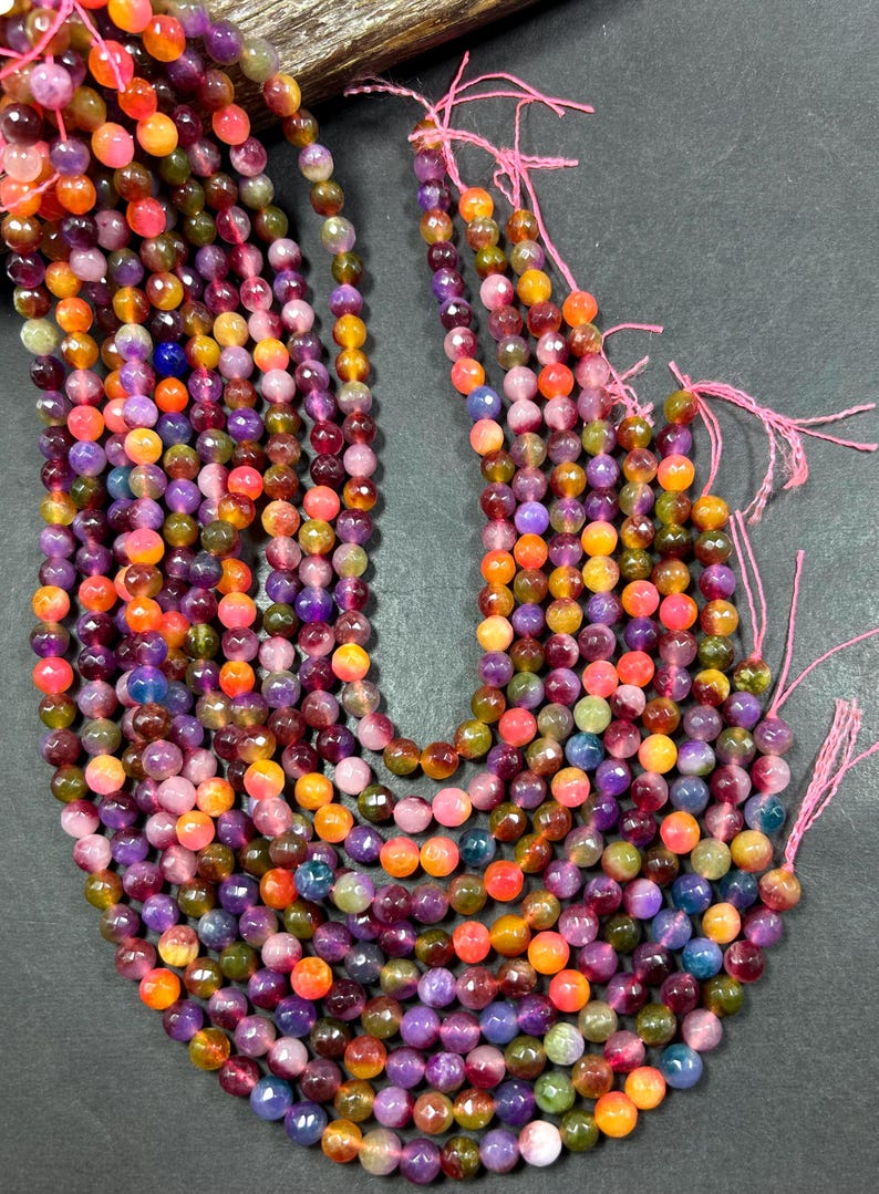 May include: A strand of multi-colored gemstone beads. The beads are round and faceted, and they are strung on a pink thread. The beads are a mix of pink, purple, orange, yellow, and green.
