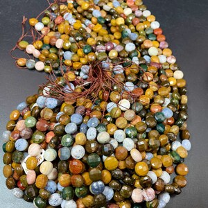 May include: A strand of multi-colored gemstone beads. The beads are round and faceted, with a variety of colors including green, brown, yellow, blue, and pink. The beads are strung on a brown thread.