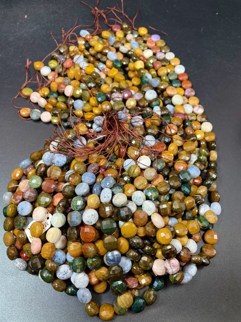 May include: A strand of multi-colored gemstone beads. The beads are round and faceted, with a variety of colors including green, brown, yellow, blue, and pink. The beads are strung on a brown thread.