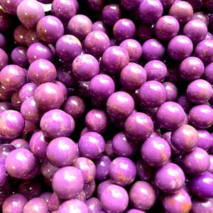 May include: A close-up shot of numerous round, polished purple beads. The beads have a smooth, glossy finish with varying shades of purple. These beads are likely used for jewellery making or other craft projects.