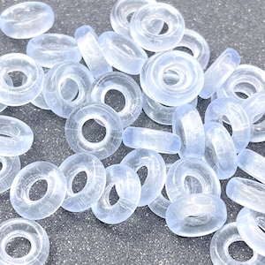 May include: A pile of clear glass donut beads. The beads are translucent and have a slightly frosted appearance.