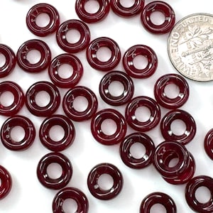 May include: A collection of small, dark red, donut-shaped beads. The beads are arranged on a white surface, with a US dime coin visible for size comparison. The beads have a glossy finish and a hole in the center.